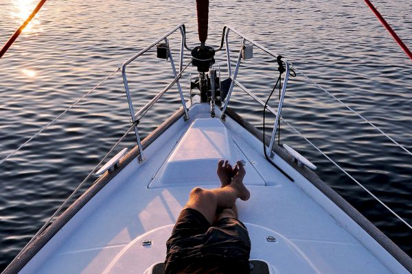 A person is relaxing on the deck of a sailboat during sunset, with calm water and distant hills in the background.