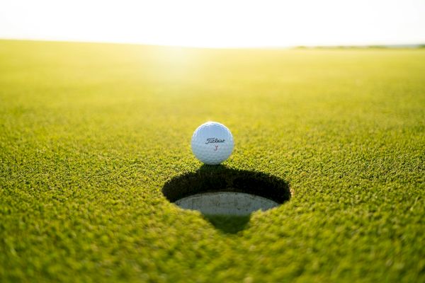 A clear golf ball sits at the edge of a golf hole on a well-manicured green under bright lighting, suggesting a golfing scenario.