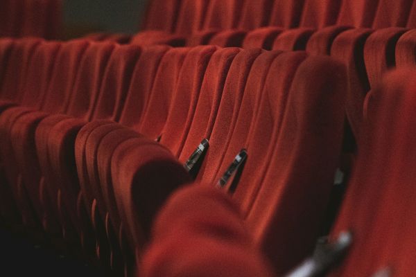 The image shows rows of empty red theater or cinema seats arranged in a dimly lit space.