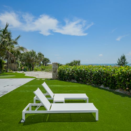 The image shows two white lounge chairs on green lawn grass, with palm trees, a house, and the ocean in the background on a sunny day.