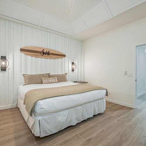 A cozy bedroom with a surfboard wall decor, beige bedding, and wood flooring, extending to a bright adjacent room.