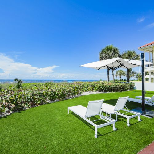 A coastal scene with a building, sun loungers on a lawn, an umbrella, and the ocean in the background under a clear blue sky.