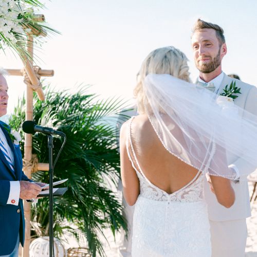 A couple is getting married on a beach, with an officiant leading the ceremony under a floral arch, surrounded by guests.