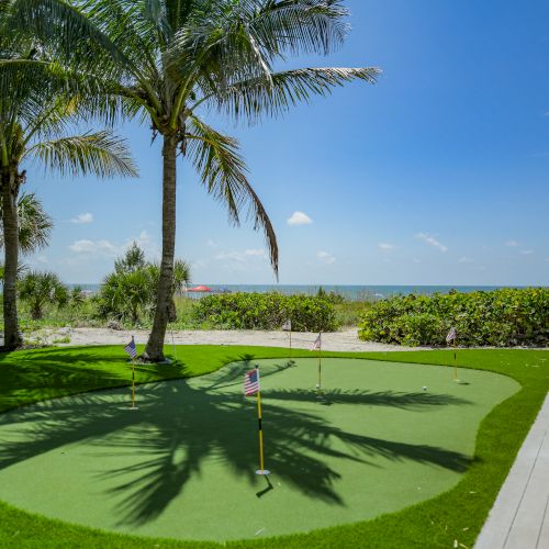 A mini-golf putting green is situated under palm trees near a beach on a sunny day. Ocean waves are visible in the background.