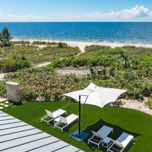 A beachfront scene with lounge chairs and an umbrella on artificial grass, overlooking a path through greenery leading to a sandy beach and the ocean.