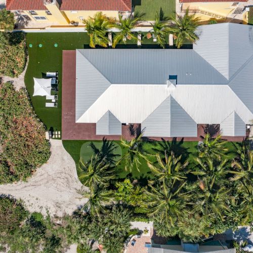 An aerial view of a building with a white roof, surrounded by lush greenery, nearby cars, and adjacent to a road.