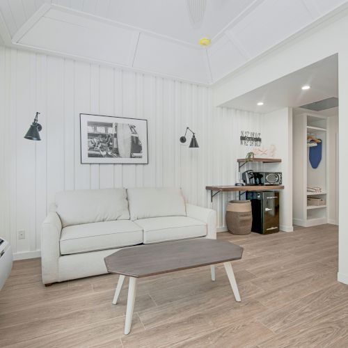 This image shows a bright living room with a white sofa, wall art, a small table, and a corner shelf with a microwave and other items, ending the sentence.