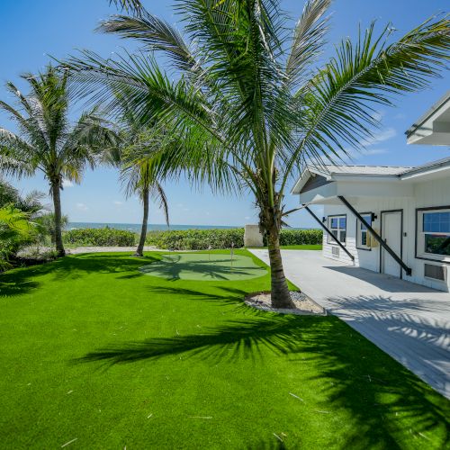 A coastal house with a deck opens up to a lush green lawn, palm trees, and a view of the ocean under a bright blue sky, ending the sentence.