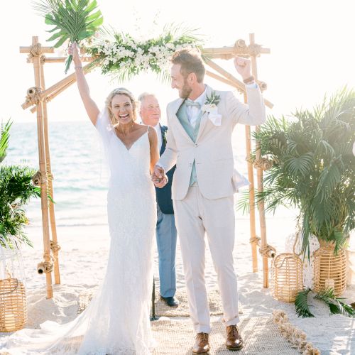 A bride and groom holding hands and raising their arms, walking down a sandy aisle under a bamboo arch, with greenery and the ocean in the background.