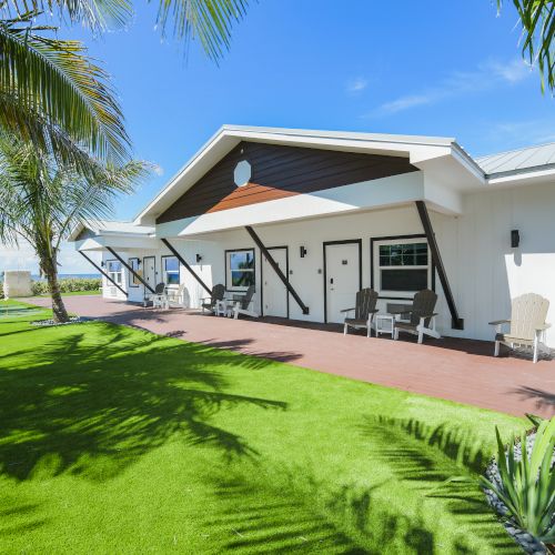 A modern one-story house with a well-maintained lawn, outdoor seating, and palm trees under a clear blue sky, ending the sentence.