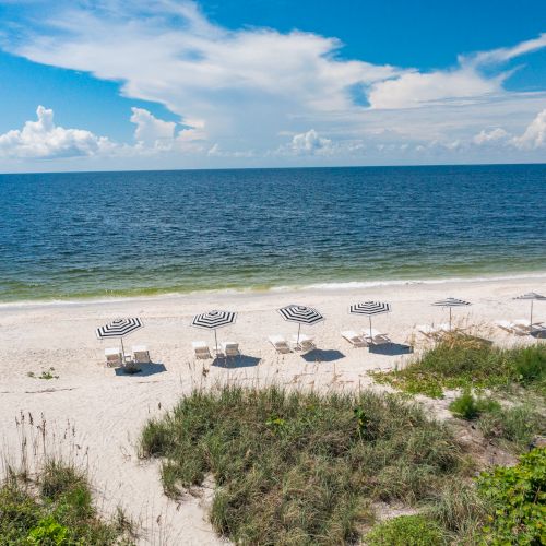 A serene beach scene with striped umbrellas, lounge chairs on white sand, a calm blue ocean, a partly cloudy sky, and some grass in the foreground.