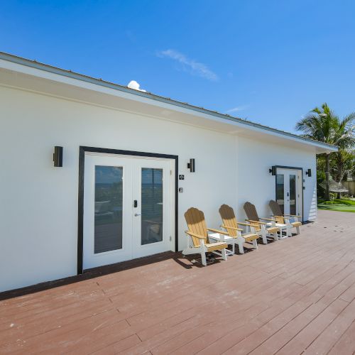 A modern white building with glass doors and a spacious wooden deck, featuring several tan deck chairs. Tropical palm trees surround the area.