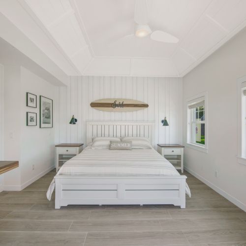 A modern bedroom with a white bed, surfboard decor above the bed, wall art, and windows providing natural light. There is also an air conditioner.