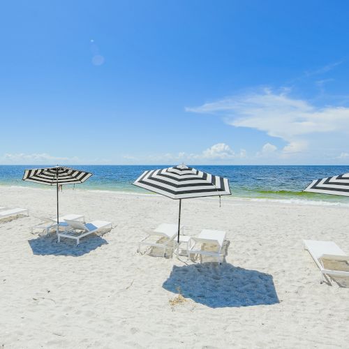 A beach scene with white sand, blue ocean, and sky. Four black-and-white striped umbrellas shade white lounge chairs placed in a row.