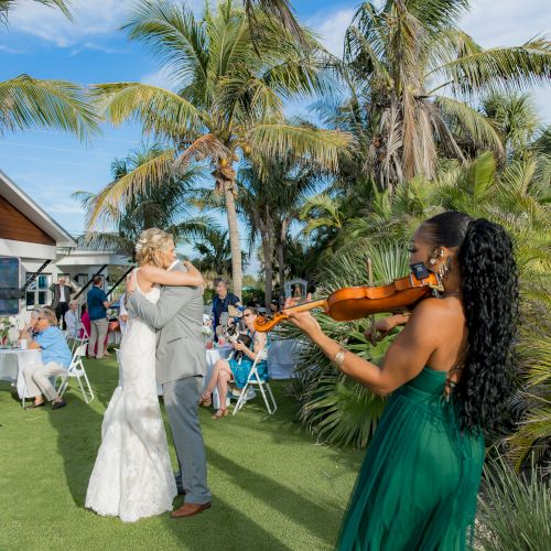 A couple is dancing at an outdoor wedding reception while a violinist in a green dress plays nearby, surrounded by palm trees and guests.