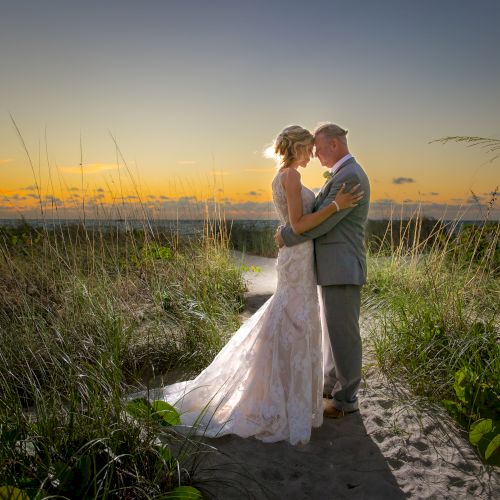 A bride and groom embrace on a sandy path at sunset, surrounded by grass and greenery, creating a romantic and serene scene.