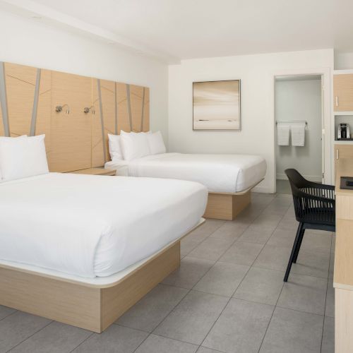 A modern hotel room with two beds, a desk, a TV, and a painting on the wall. The decor is minimalist with light wooden furniture and neutral tones.