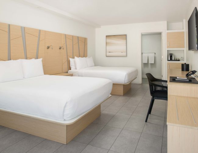 A modern hotel room with two beds, a desk, a TV, and a painting on the wall. The decor is minimalist with light wooden furniture and neutral tones.