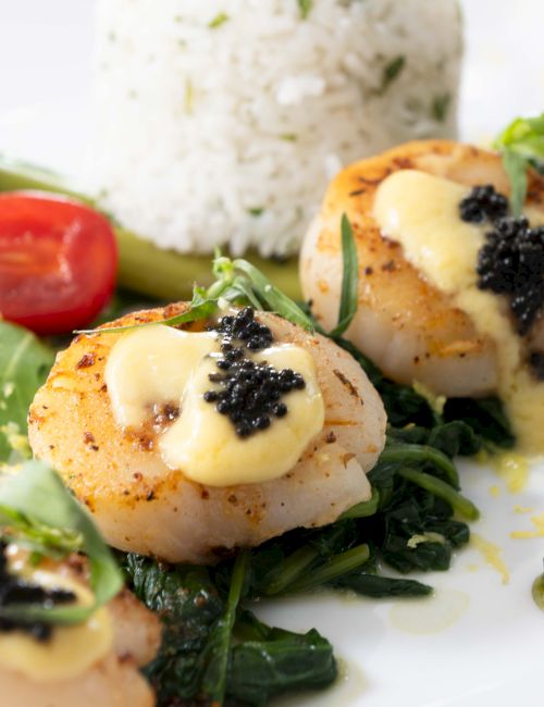 Scallops presented on a bed of greens, topped with a creamy sauce and caviar, accompanied by rice, asparagus, and a cherry tomato.