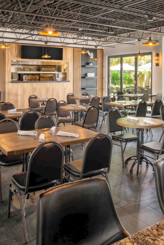 The image shows a modern, empty restaurant with wooden tables, black chairs, large windows, and a visible kitchen area at the far end.