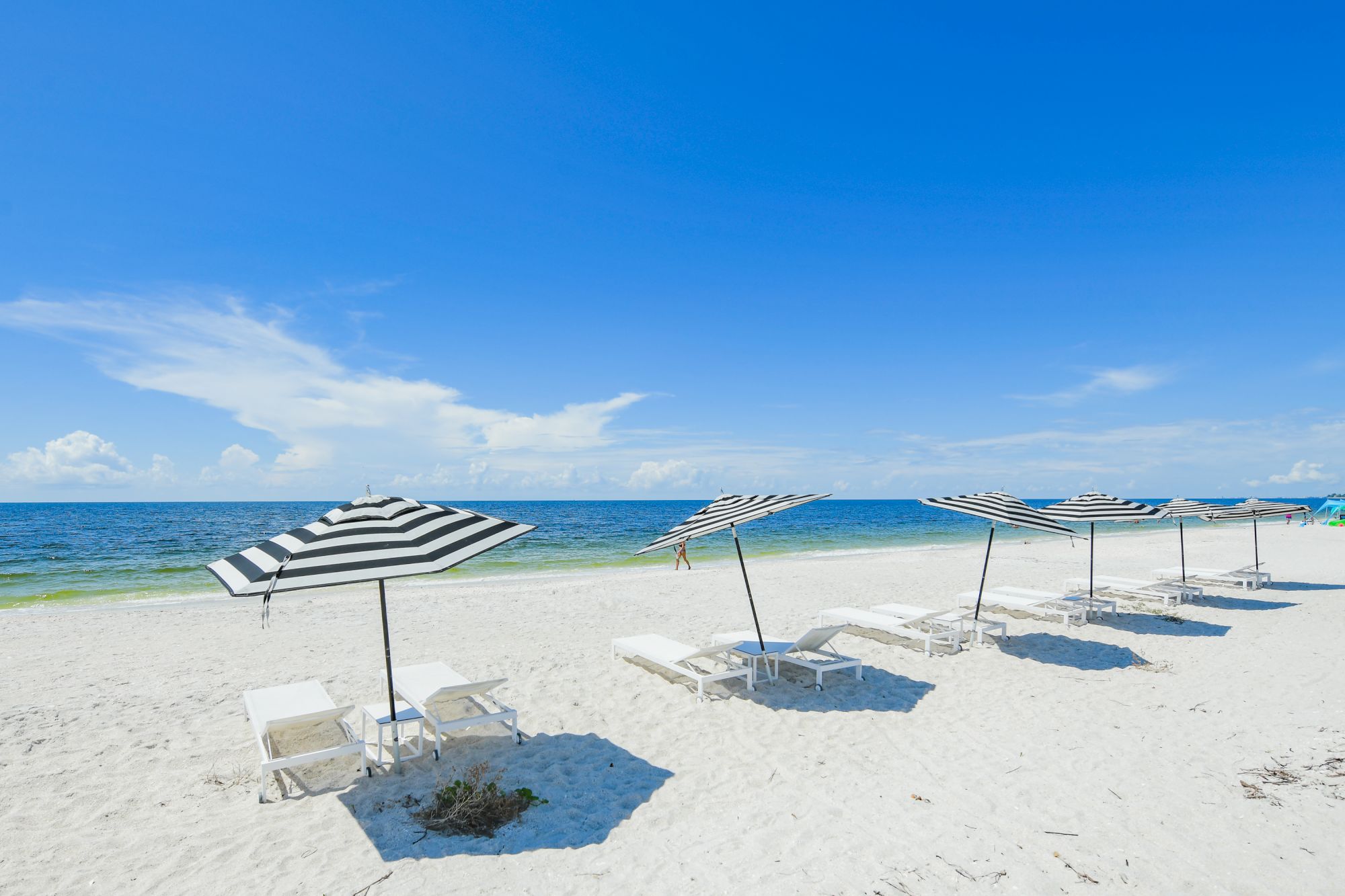 White sandy beach with empty lounge chairs and striped umbrellas, facing a calm blue ocean under a clear sky.