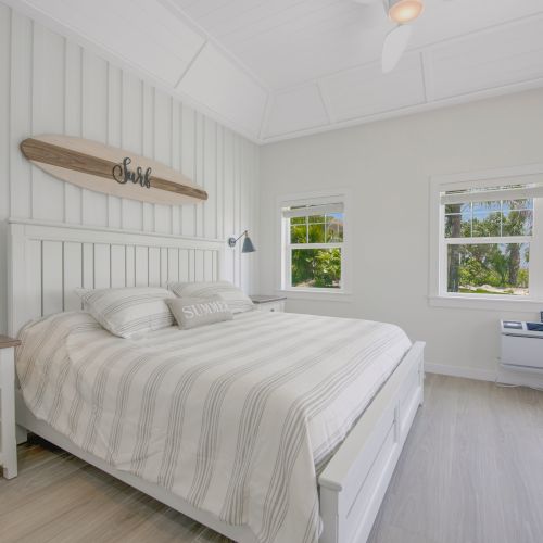 A bright bedroom with a large bed, striped bedding, two bedside tables, wall sconces, windows with a garden view, and a surfboard wall decor, always ending.