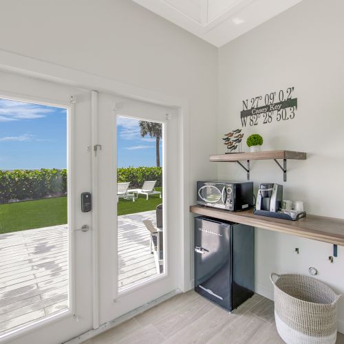 A bright room with a coffee maker, microwave, mini-fridge, and basket, leads to a deck and lawn with patio furniture, under a clear blue sky.