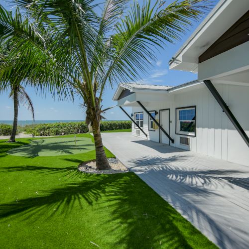 A modern house with white siding is bordered by a wooden deck, lush green lawn, and tropical palm trees, with the ocean visible in the background.