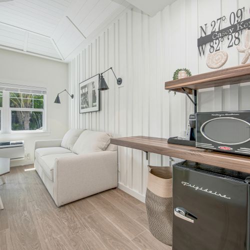 A cozy, modern living space with a white couch, wall art, a coffee table, and a kitchenette with a microwave and mini fridge on a wooden counter.