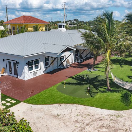 A white house with a metal roof has a wooden deck, hammock, and putting green surrounded by palm trees and greenery, under a partly cloudy sky.