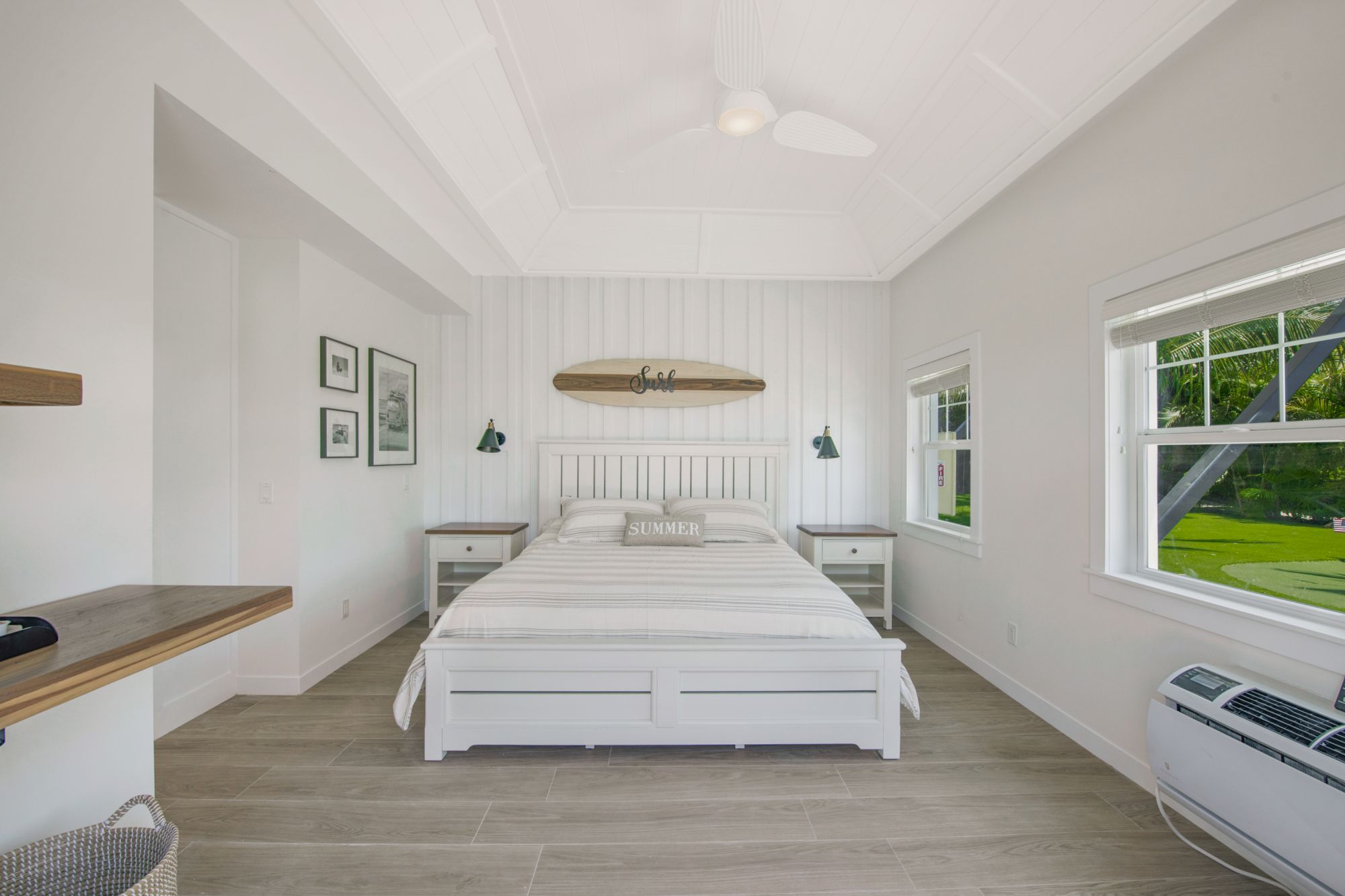 A bright bedroom with a white bed, surfboard above, bedside tables, framed photos, large windows, a desk, and a wicker basket on the floor.