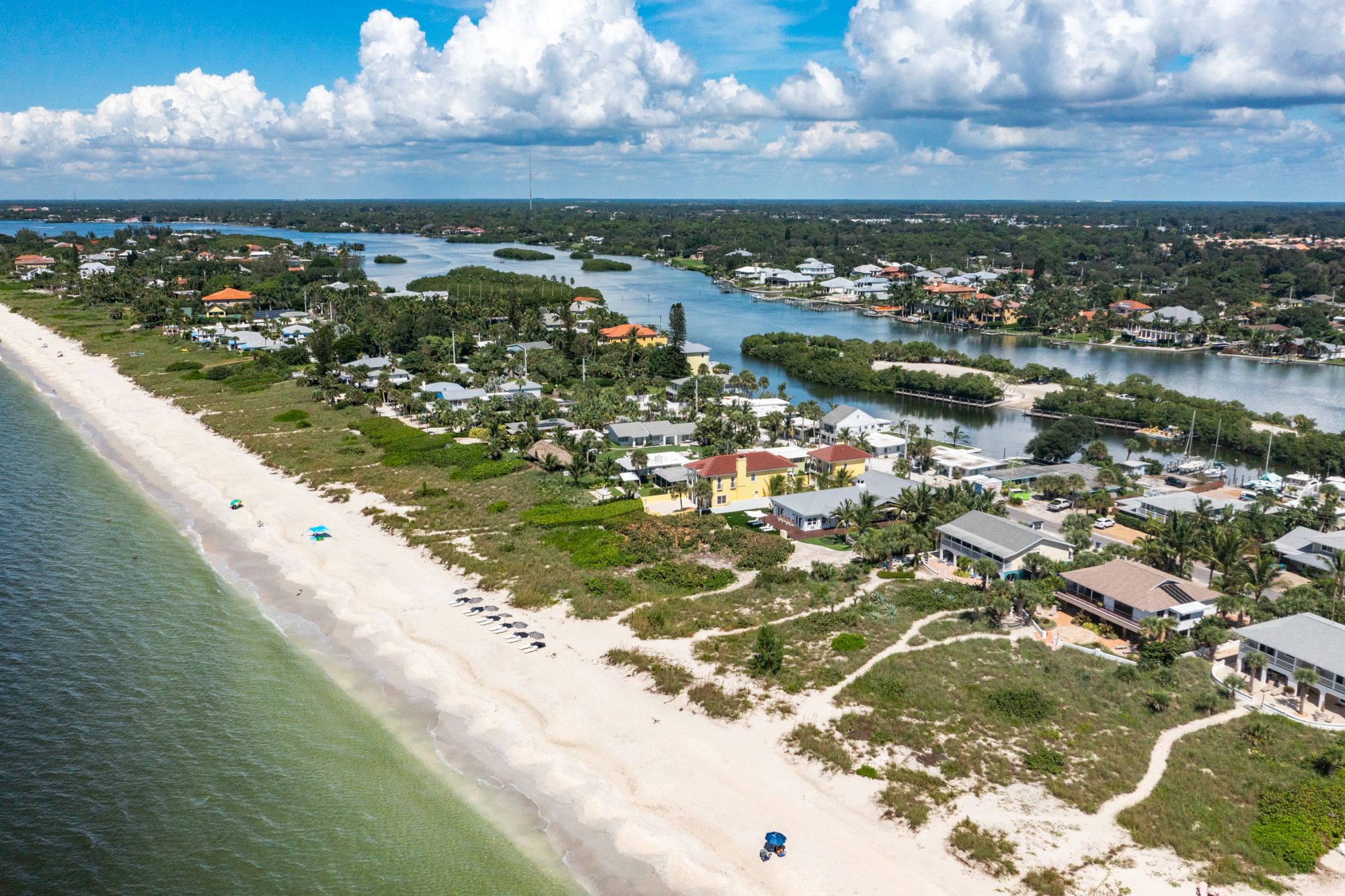 A scenic aerial view of a beachfront community with houses, lush greenery, sandy shores, and ocean waves under a partly cloudy sky.