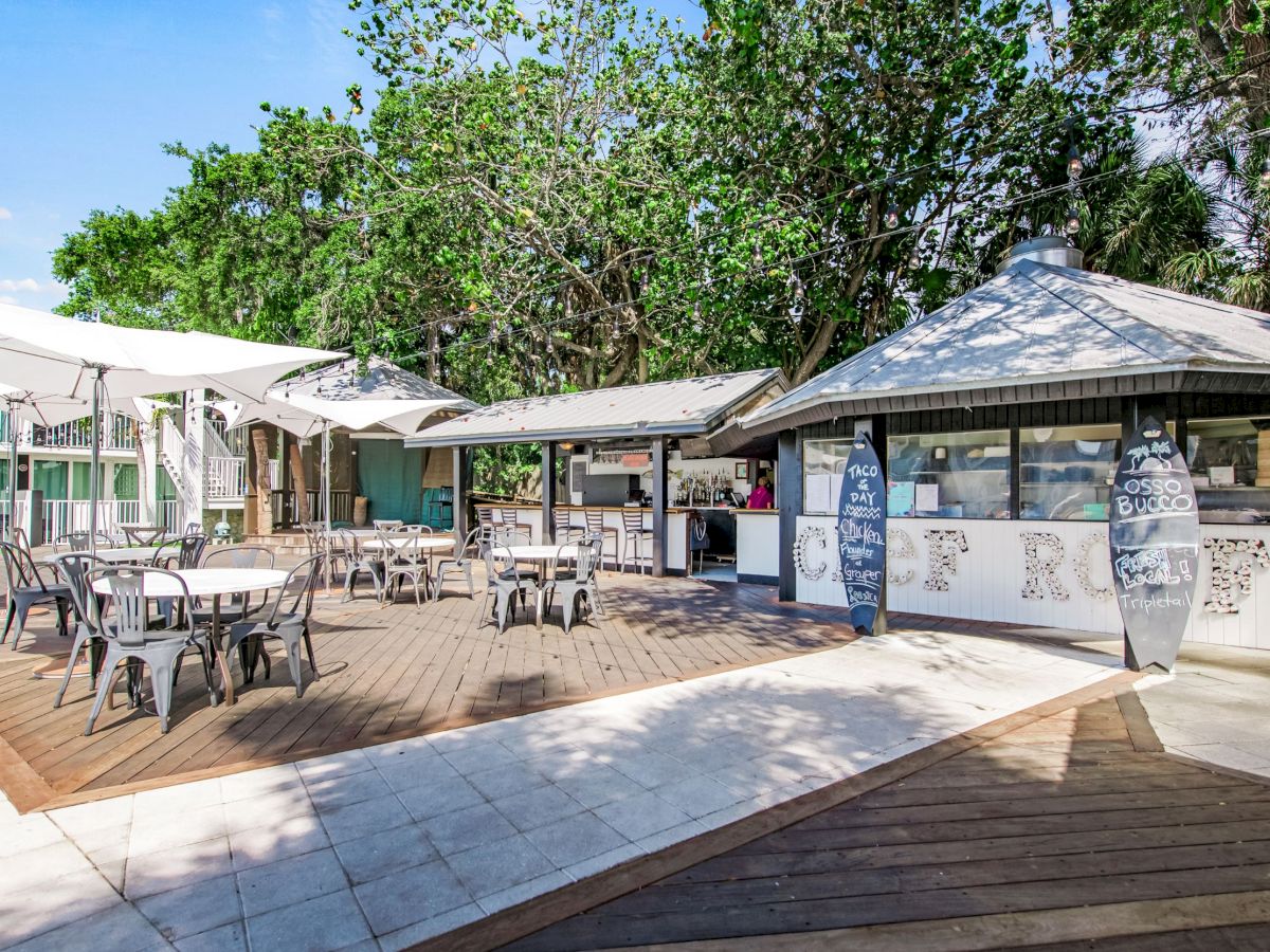 An outdoor cafe with tables and chairs, shaded by umbrellas, surrounded by trees. There are buildings in the background and surfboards.