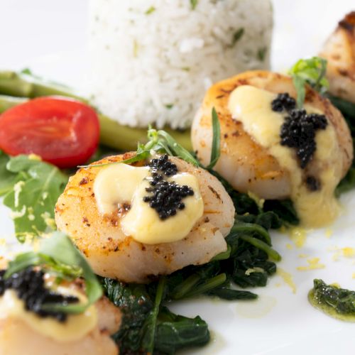 The image shows a gourmet dish with seared scallops topped with a creamy sauce and caviar, served with greens, asparagus, a cherry tomato, and rice.