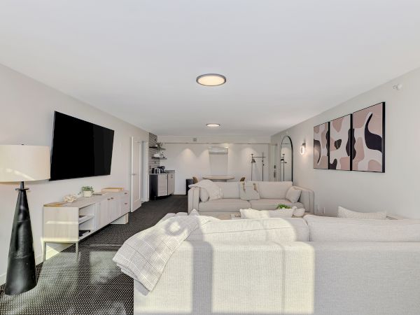 The image shows a modern living room with sofas, a wall-mounted TV, large artwork, and minimalistic decor in a neutral color palette.