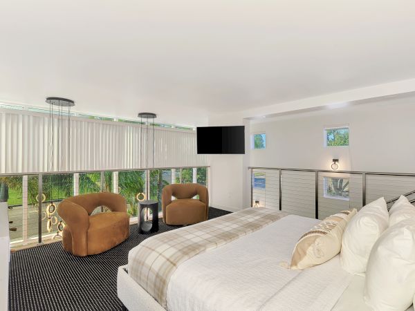 A modern bedroom with a large bed, two armchairs, a wall-mounted TV, and large windows with blinds, creating a cozy and inviting atmosphere.