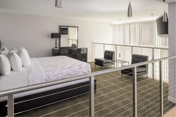 A modern bedroom with a large bed, desk, two lounge chairs, TV, mirror, and vertical blinds, featuring a minimalist design and neutral tones.