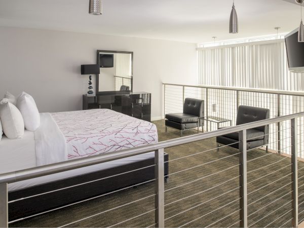 A modern bedroom with a large bed, desk, two lounge chairs, TV, mirror, and vertical blinds, featuring a minimalist design and neutral tones.