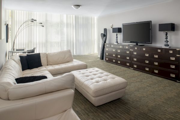 A modern living room with white sectional sofa, ottoman, large TV on a dark cabinet, floor lamp, and vertical blinds covering large windows.