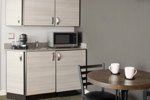 A small kitchen area with cabinets, a microwave, a coffee maker, and a round table with two chairs and two white mugs on it, ending the sentence.