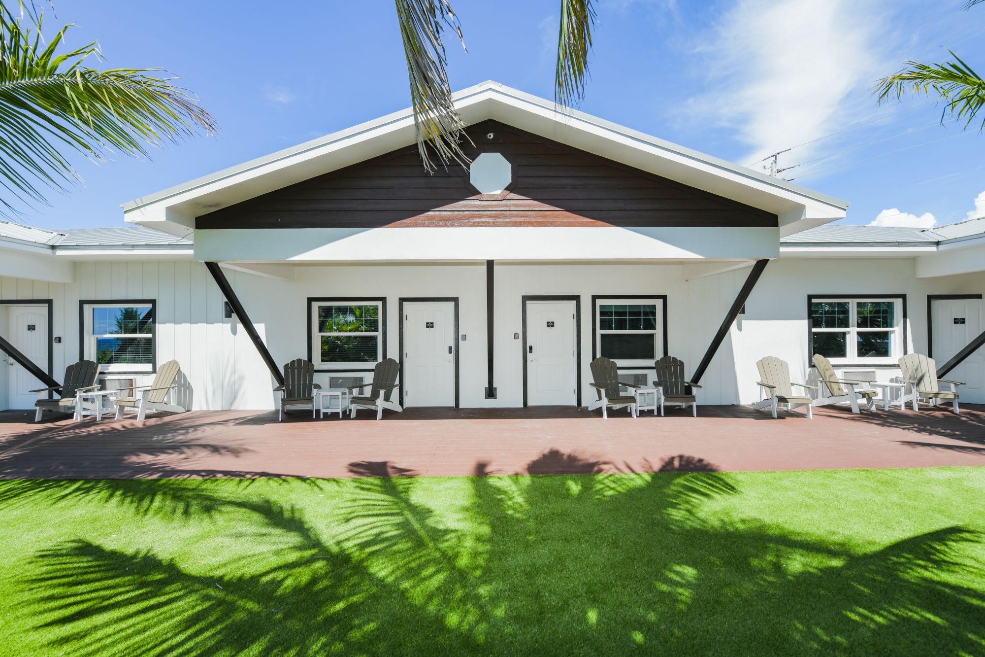 A building with a white exterior, three doors, patio chairs, tables, and lush green grass under a clear sky with palm tree shadows ends the sentence.