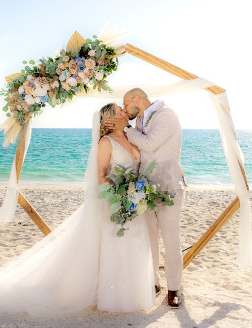 A couple kisses at their beach wedding under a hexagonal floral arch, with the ocean in the background and sand beneath them.