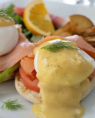 The image shows Eggs Benedict with poached eggs, hollandaise sauce, asparagus, smoked salmon, tomato on an English muffin, and roasted potatoes with orange slice.