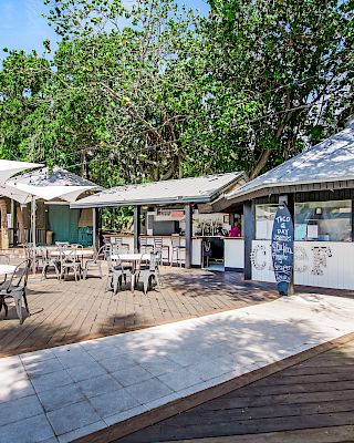 Outdoor café with tables, chairs, and surfboards. It has shaded areas and trees in the background, offering a relaxed atmosphere.