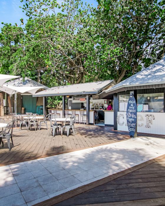 Outdoor caf&eacute; with tables, chairs, and surfboards. It has shaded areas and trees in the background, offering a relaxed atmosphere.