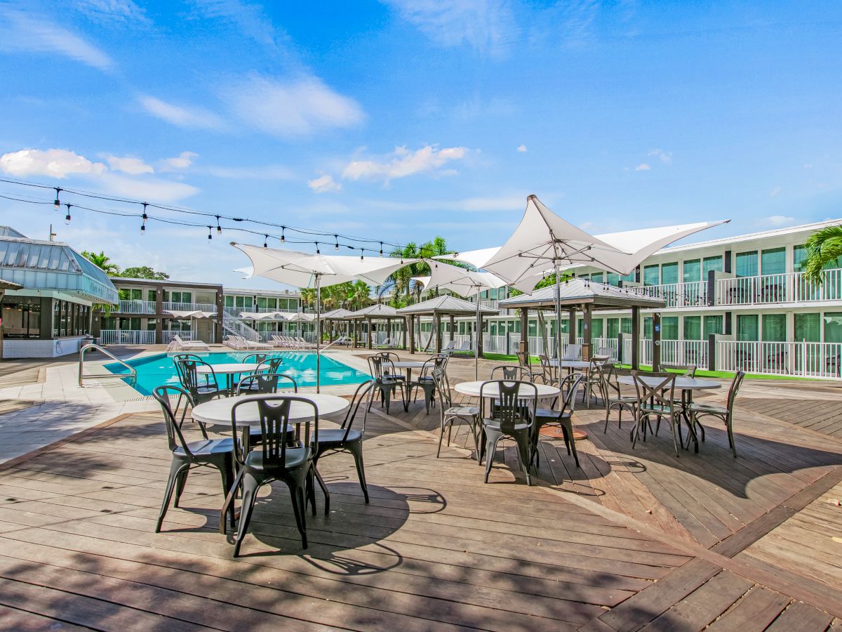 An outdoor pool area with tables, chairs, and umbrellas on a wooden deck is adjacent to multi-story buildings. Strings of lights hang overhead.