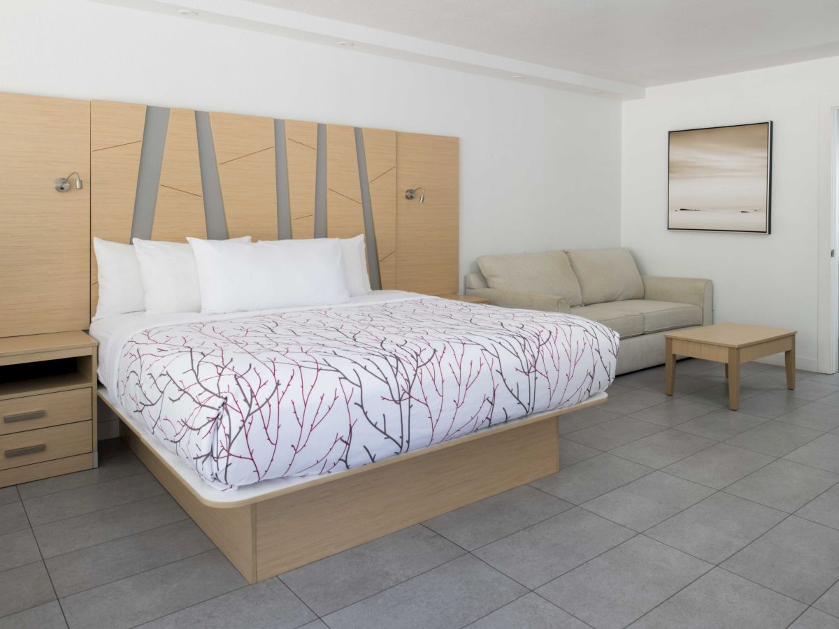 A modern hotel room with a bed, gray sofa, wooden nightstands, coffee table, and abstract art on the wall, featuring a simple, clean design.