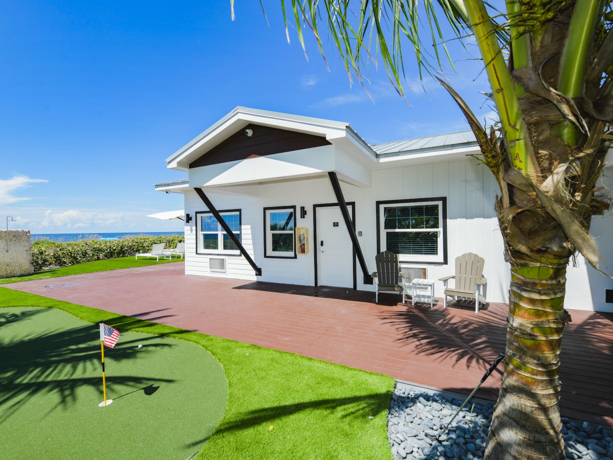 A modern white house with a wooden deck, palm tree, and a small putting green in the yard on a sunny day with blue sky in the background.