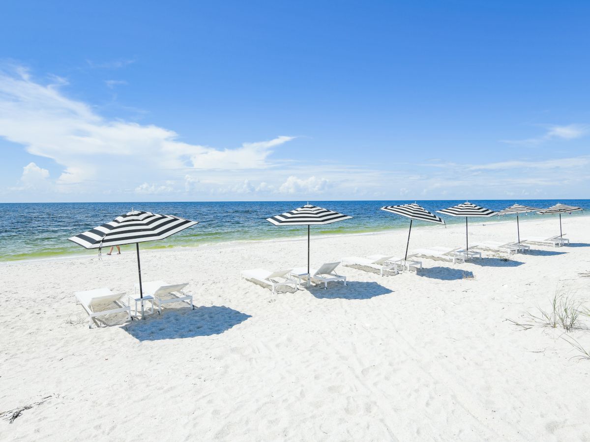 A pristine beach with white sand, featuring six black-and-white striped umbrellas and lounge chairs, all facing the calm blue ocean waters.