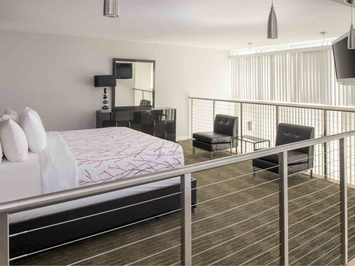 A modern loft bedroom featuring a bed, desk with mirror, two chairs, a TV, and hanging lights, all with a minimalist design and neutral colors.